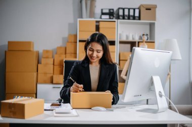 Girişimci Asyalı kadın çevrimiçi alışveriş için tablet ve dizüstü bilgisayarda paketler hazırlıyor ve çevrimiçi siparişleri kontrol ediyor.