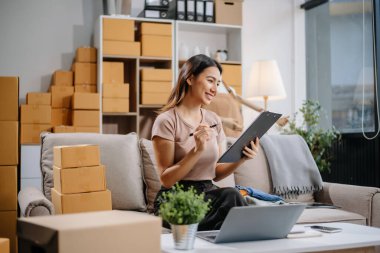 Girişimci Asyalı kadın çevrimiçi alışveriş için tablet ve dizüstü bilgisayarda paketler hazırlıyor ve çevrimiçi siparişleri kontrol ediyor.