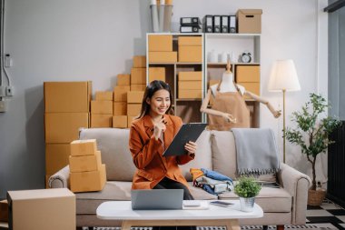 Girişimci Asyalı kadın çevrimiçi alışveriş için tablet ve dizüstü bilgisayarda paketler hazırlıyor ve çevrimiçi siparişleri kontrol ediyor.