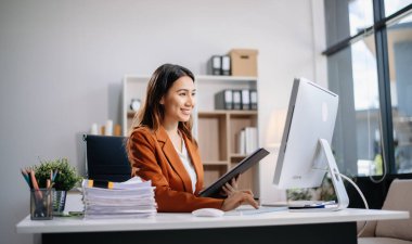 Genç Asyalı kadın, gülümsüyor ve ofis ortamında masasında çalışıyor. Bir iş elbisesi giyiyor ve dizüstü bilgisayar kullanırken elinde belgeler tutuyor.