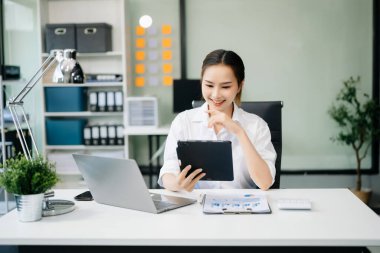 Çalışan not defteri, tablet ve dizüstü bilgisayarla çalışan genç Asyalı iş kadını akıllı telefon, tablet ve dizüstü bilgisayarla konuşuyor. Video arama vergisi, rapor, muhasebe 