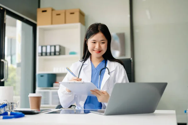 Confident young Asian female doctor in white medical uniform sit at desk working on computer. Smiling use laptop write in medical journal in clinic or hospital.