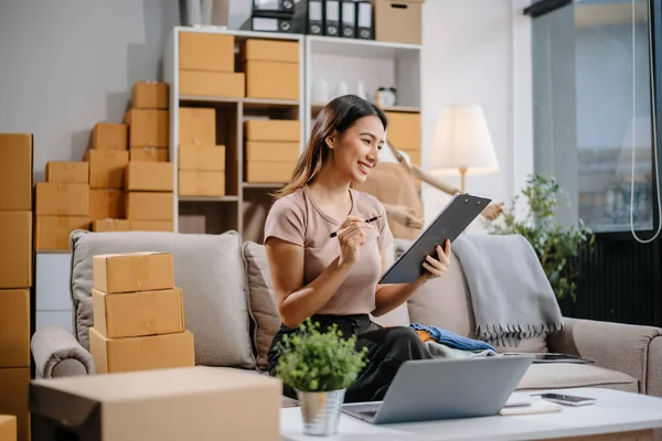 Entrepreneurial Asian woman prepares parcels and checks online orders on tablet and laptop for delivery online shopping and small business success in office