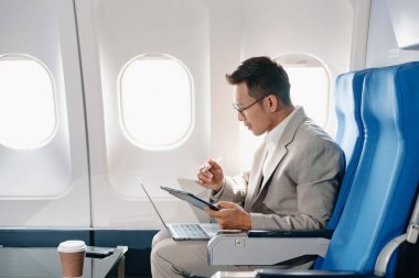 Genç Asyalı yönetici, dijital tablet, dizüstü bilgisayar ve akıllı telefondan birden fazla iş yaparak birinci sınıfta başarılı oluyor. Stil içinde seyahat et, zarafetle çalış.