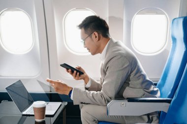 Genç Asyalı yönetici, dijital tablet, dizüstü bilgisayar ve akıllı telefondan birden fazla iş yaparak birinci sınıfta başarılı oluyor. Stil içinde seyahat et, zarafetle çalış.