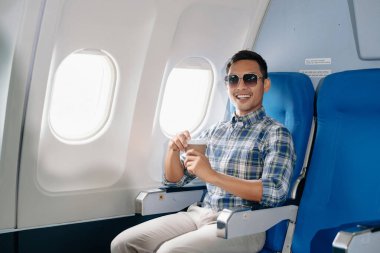 Çekici Asyalı erkek uçak yolcusu dizüstü bilgisayar ve tabletle kablosuz bağlantıyı kullanarak rahat koltukta oturuyor. Stil içinde seyahat et, zarafetle çalış.