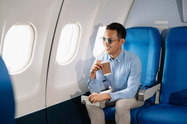 Çekici Asyalı erkek uçak yolcusu dizüstü bilgisayar ve tabletle kablosuz bağlantıyı kullanarak rahat koltukta oturuyor. Stil içinde seyahat et, zarafetle çalış.