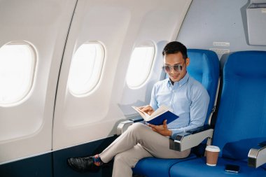 Çekici Asyalı erkek uçak yolcusu dizüstü bilgisayar ve tabletle kablosuz bağlantıyı kullanarak rahat koltukta oturuyor. Stil içinde seyahat et, zarafetle çalış.