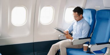 Çekici Asyalı erkek uçak yolcusu dizüstü bilgisayar ve tabletle kablosuz bağlantıyı kullanarak rahat koltukta oturuyor. Stil içinde seyahat et, zarafetle çalış.