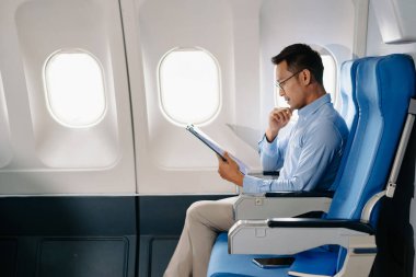 Çekici Asyalı erkek uçak yolcusu dizüstü bilgisayar ve tabletle kablosuz bağlantıyı kullanarak rahat koltukta oturuyor. Stil içinde seyahat et, zarafetle çalış.