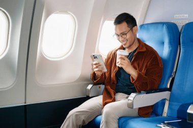 Çekici Asyalı erkek uçak yolcusu dizüstü bilgisayar ve tabletle kablosuz bağlantıyı kullanarak rahat koltukta oturuyor. Stil içinde seyahat et, zarafetle çalış.
