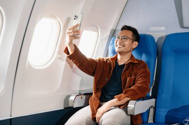 Çekici Asyalı erkek uçak yolcusu dizüstü bilgisayar ve tabletle kablosuz bağlantıyı kullanarak rahat koltukta oturuyor. Stil içinde seyahat et, zarafetle çalış.