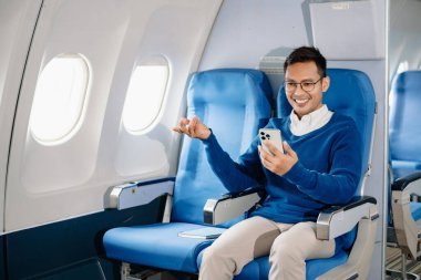 Çekici Asyalı erkek uçak yolcusu dizüstü bilgisayar ve tabletle kablosuz bağlantıyı kullanarak rahat koltukta oturuyor. Stil içinde seyahat et, zarafetle çalış.