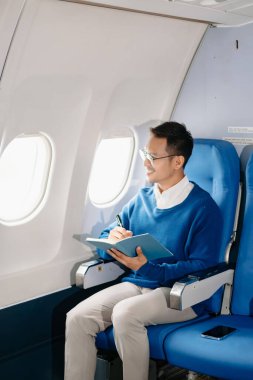 Çekici Asyalı erkek uçak yolcusu dizüstü bilgisayar ve tabletle kablosuz bağlantıyı kullanarak rahat koltukta oturuyor. Stil içinde seyahat et, zarafetle çalış.