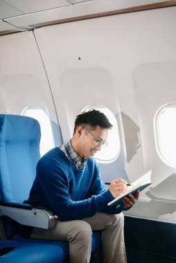 Çekici Asyalı erkek uçak yolcusu dizüstü bilgisayar ve tabletle kablosuz bağlantıyı kullanarak rahat koltukta oturuyor. Stil içinde seyahat et, zarafetle çalış.