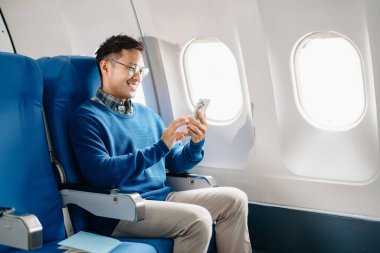 Çekici Asyalı erkek uçak yolcusu dizüstü bilgisayar ve tabletle kablosuz bağlantıyı kullanarak rahat koltukta oturuyor. Stil içinde seyahat et, zarafetle çalış.