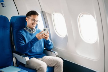 Çekici Asyalı erkek uçak yolcusu dizüstü bilgisayar ve tabletle kablosuz bağlantıyı kullanarak rahat koltukta oturuyor. Stil içinde seyahat et, zarafetle çalış.