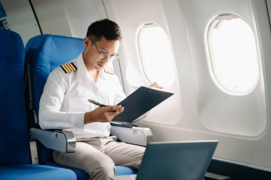 Asyalı kendinden emin üniformalı erkek pilot kalkış sırasında ve uçuş planlarını kontrol ederken yolcu koltuğuna yaslanıyor.