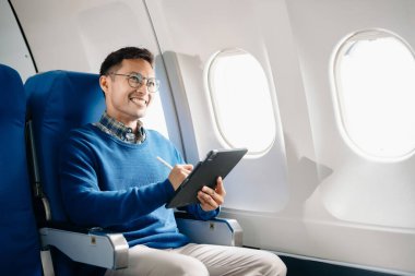 Çekici Asyalı erkek uçak yolcusu dizüstü bilgisayar ve tabletle kablosuz bağlantıyı kullanarak rahat koltukta oturuyor. Stil içinde seyahat et, zarafetle çalış.