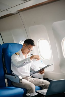 Asyalı kendinden emin üniformalı erkek pilot kalkış sırasında ve uçuş planlarını kontrol ederken yolcu koltuğuna yaslanıyor.