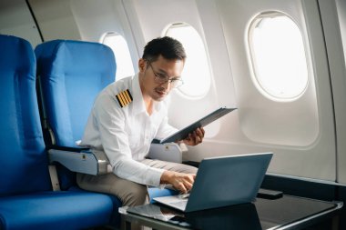 Asyalı kendinden emin üniformalı erkek pilot kalkış sırasında ve uçuş planlarını kontrol ederken yolcu koltuğuna yaslanıyor.
