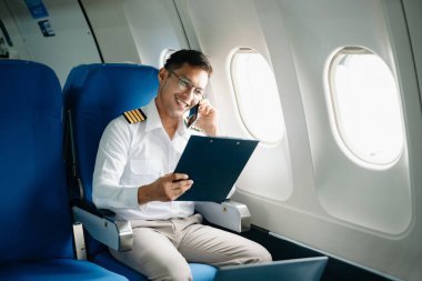 Asyalı kendinden emin üniformalı erkek pilot kalkış sırasında ve uçuş planlarını kontrol ederken yolcu koltuğuna yaslanıyor.