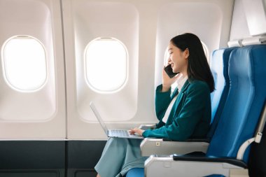 Çekici Asyalı kadın uçak yolcusu dizüstü bilgisayar ve tabletle kablosuz bağlantı kullanarak rahat koltukta oturuyor. Stil ve iş seyahatleri. 
