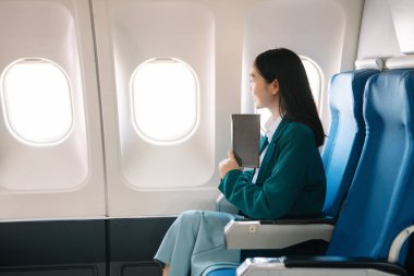 Çekici Asyalı kadın uçak yolcusu dizüstü bilgisayar ve tabletle kablosuz bağlantı kullanarak rahat koltukta oturuyor. Stil ve iş seyahatleri. 