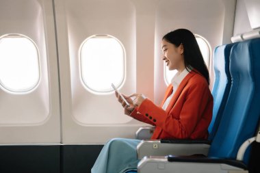 Çekici Asyalı kadın uçak yolcusu dizüstü bilgisayar ve tabletle kablosuz bağlantı kullanarak rahat koltukta oturuyor. Stil ve iş seyahatleri. 