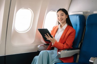 Çekici Asyalı kadın uçak yolcusu dizüstü bilgisayar ve tabletle kablosuz bağlantı kullanarak rahat koltukta oturuyor. Stil ve iş seyahatleri. 