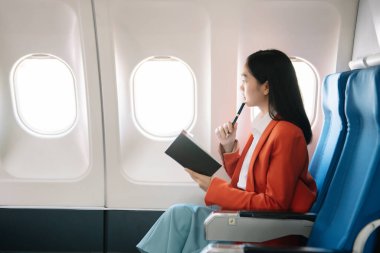 Çekici Asyalı kadın uçak yolcusu dizüstü bilgisayar ve tabletle kablosuz bağlantı kullanarak rahat koltukta oturuyor. Stil ve iş seyahatleri. 