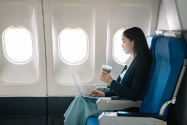Çekici Asyalı kadın uçak yolcusu dizüstü bilgisayar ve tabletle kablosuz bağlantı kullanarak rahat koltukta oturuyor. Stil ve iş seyahatleri. 