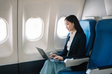 Çekici Asyalı kadın uçak yolcusu dizüstü bilgisayar ve tabletle kablosuz bağlantı kullanarak rahat koltukta oturuyor. Stil ve iş seyahatleri. 