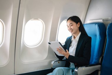 Çekici Asyalı kadın uçak yolcusu dizüstü bilgisayar ve tabletle kablosuz bağlantı kullanarak rahat koltukta oturuyor. Stil ve iş seyahatleri. 