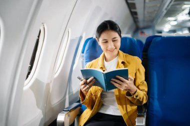 Çekici Asyalı kadın uçak yolcusu dizüstü bilgisayar ve tabletle kablosuz bağlantı kullanarak rahat koltukta oturuyor. Stil içinde seyahat et, zarafetle çalış..