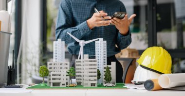 Ofisteki masada dizüstü bilgisayar ve tabletle çalışan genç bir emlakçı ve yanında da küçük bir ev..