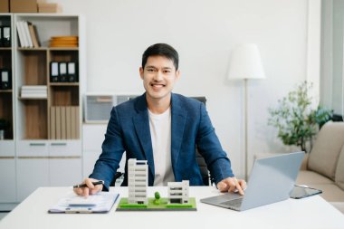 Ofisteki masada dizüstü bilgisayar ve tabletle çalışan genç bir emlakçı ve yanında da küçük bir ev..