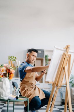 Asyalı erkek ressam sanat atölyesinde resim yapıyor, resim malzemeleri, yağ boyama sehpası, iki tuval sehpası, resim stüdyosunda veya oturma odasında fırçalı yaratıcı bir alan.