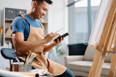 Asyalı erkek ressam sanat atölyesinde resim yapıyor, resim malzemeleri, yağ boyama sehpası, iki tuval sehpası, resim stüdyosunda veya oturma odasında fırçalı yaratıcı bir alan.
