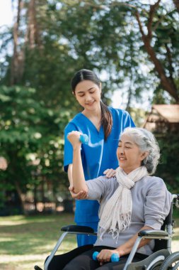 Asyalı fizyoterapist, yaşlı bir kadının egzersiz sırasında kollarını esnetmesine yardım ediyor. Hasta sırtı problemli bir halterle, ellerindeki dambılla. Huzurevi hastane bahçesi konsepti