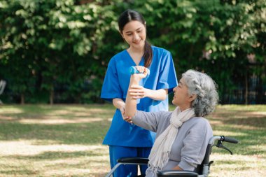 Asyalı fizyoterapist, yaşlı bir kadının egzersiz sırasında kollarını esnetmesine yardım ediyor. Hasta sırtı problemli bir halterle, ellerindeki dambılla. Huzurevi hastane bahçesi konsepti