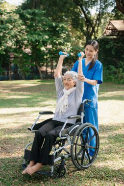 Asyalı fizyoterapist, yaşlı bir kadının egzersiz sırasında kollarını esnetmesine yardım ediyor. Hasta sırtı problemli bir halterle, ellerindeki dambılla. Huzurevi hastane bahçesi konsepti