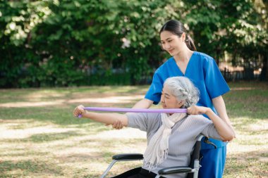 Asyalı fizyoterapist, yaşlı bir kadının egzersiz sırasında kollarını esnetmesine yardım ediyor. Hasta sırtı problemli bir halterle, ellerindeki dambılla. Huzurevi hastane bahçesi konsepti