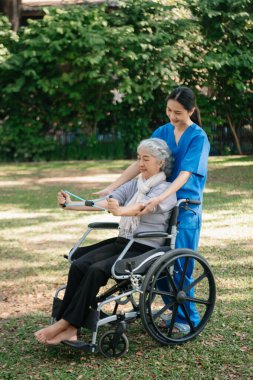 Asyalı fizyoterapist, yaşlı bir kadının egzersiz sırasında kollarını esnetmesine yardım ediyor. Hasta sırtı problemli bir halterle, ellerindeki dambılla. Huzurevi hastane bahçesi konsepti