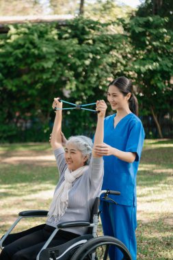 Asyalı fizyoterapist, yaşlı bir kadının egzersiz sırasında kollarını esnetmesine yardım ediyor. Hasta sırtı problemli bir halterle, ellerindeki dambılla. Huzurevi hastane bahçesi konsepti