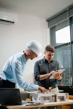 Mimaride profesyonel işbirliği. İki mühendis, dizüstü bilgisayarlar, tabletler ve ölçekli modeller gibi araçlar kullanarak inşaat planları üzerinde çalışıyor. Takım çalışması için mükemmel bir tasvir. 