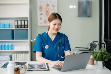 Beyaz üniformalı, kendine güvenen Asyalı kadın doktor masa başında bilgisayar üzerinde çalışıyor. Klinikteki tıp dergisine gülümseyen dizüstü bilgisayar yazıları.