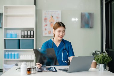 Beyaz üniformalı, kendine güvenen Asyalı kadın doktor masa başında bilgisayar üzerinde çalışıyor. Klinikteki tıp dergisine gülümseyen dizüstü bilgisayar yazıları.