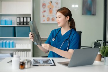 Beyaz üniformalı, kendine güvenen Asyalı kadın doktor masa başında bilgisayar üzerinde çalışıyor. Klinikteki tıp dergisine gülümseyen dizüstü bilgisayar yazıları.