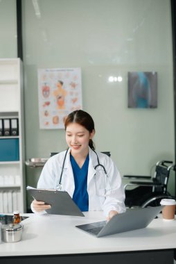 Beyaz üniformalı, kendine güvenen Asyalı kadın doktor masa başında bilgisayar üzerinde çalışıyor. Klinikteki tıp dergisine gülümseyen dizüstü bilgisayar yazıları.